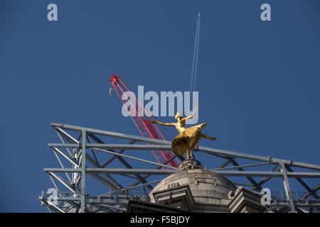 London, UK. 01st Nov, 2015. The gold statue of prima ballerina Anna Pavlova strikes an elegant pose on top of the Victoria Palace Theatre, Victoria, London, with construction of the new Victoria Centre and vapor trails of a jet airliner extending behind at diagonal angles against a blue sky Credit:  On Sight Photographic/Alamy Live News Stock Photo