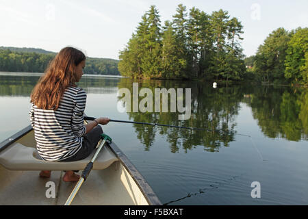 Young girl fishing from a canoe on a calm lake with trees in the background Stock Photo