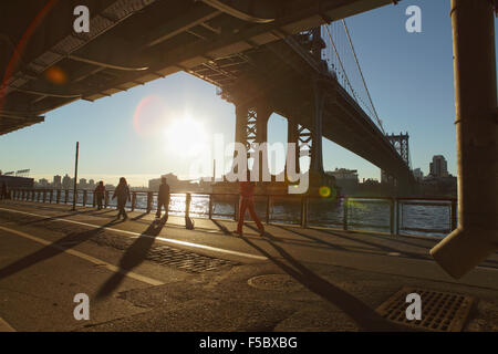 Four New Yorkers walk up the East River Esplanade under the Manhattan Bridge as the sunrise flares into the camera lens Stock Photo