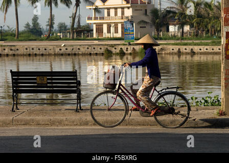 Cyclists woman on the promenade at the Thu Bon River in Hoi An Vietnam. Typical vietnamise woman with hat. Stock Photo