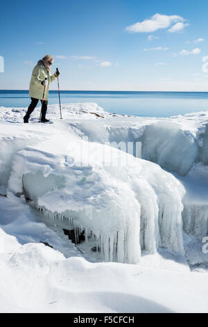 Senior adult Caucasian woman carefully walks with ski poles over icy snow covered rocks along the frozen shore of Lake Superior Stock Photo