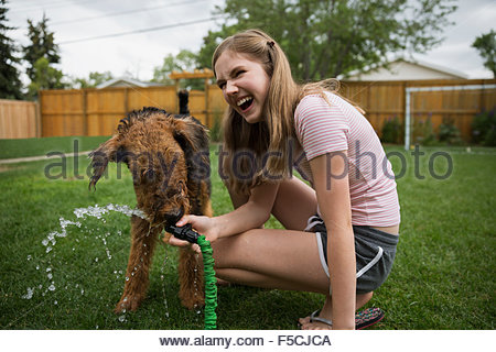 Laughing girl holding hose for dog drinking water