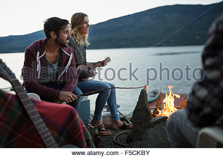 Young friends roasting marshmallows playing ukulele lakeside campfire