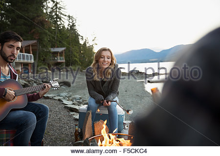 Young friends roasting marshmallows playing guitar lakeside campfire