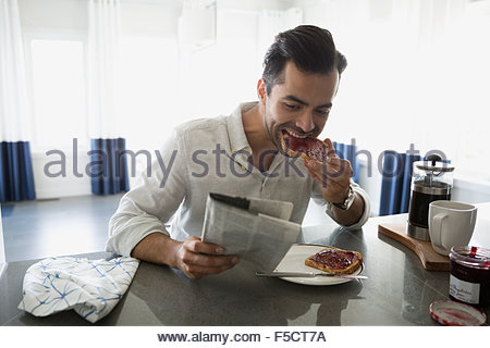 Man reading newspaper and eating toast in kitchen