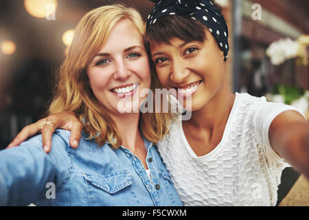 Two young women best friends sitting arm in arm with their faces close together smiling at the camera, multiethnic couple Stock Photo
