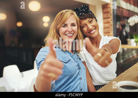 Two happy young female friends giving a thumbs up gesture of approval and success as they sit arm in arm in a cafeteria enjoying