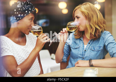 Two attractive young women meeting up in a pub for a glass of white wine sitting at a counter smiling at each other as they sip Stock Photo