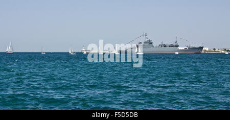 Big military ship and many little white sailing boats around in sea. Recorded in town Sevastopol in Crimea on Black sea. Stock Photo