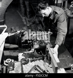 1950s historical, male worker at work bench wearing an army jacket using a machine to cut a piece of wood. Stock Photo