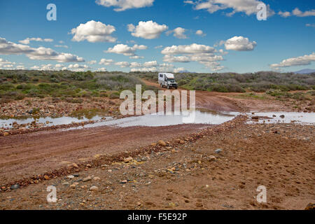 Four wheel drive campervan / motorhome about to drive through water at creek crossing on Australian outback road hemmed by low vegetation after rain Stock Photo