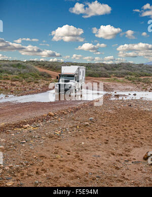 Four wheel drive campervan / motorhome driving through water of creek in outback Australia on red dirt road hemmed by low vegetation after rain Stock Photo