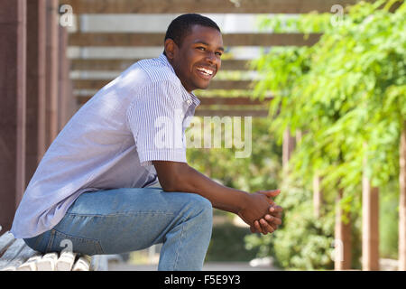 Close up portrait of a smiling relaxed young black guy sitting on bench in the park Stock Photo