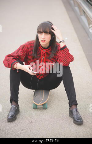 Young beautiful caucasian woman seated on a skate using a smartphone listening to music overlooking - technology, social network, communication concept Stock Photo