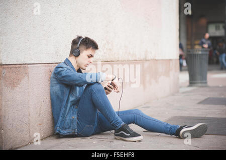 young handsome alternative dark man listening to music sitting on a sidewalk with headphones and smartphone connected wireless, Stock Photo