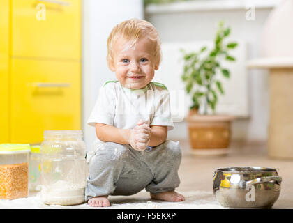 Playful child toddler with face soiled flour. Little boy surrounded kitchenware and foodstuffs Stock Photo