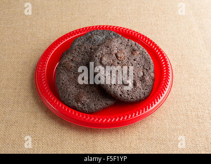 Three freshly baked chocolate chip crispy brownie cookies on a red plate atop a burlap tablecloth illuminated with natural light Stock Photo