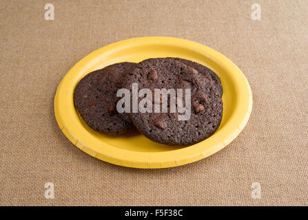 Three freshly baked chocolate chip crispy brownie cookies on a yellow paper plate atop a burlap tablecloth Stock Photo
