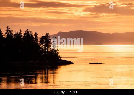 Mountains and trees in Alaska silhouetted by the setting sun with sunlight reflecting off the water. Stock Photo