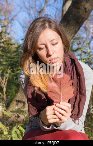 Sad girl holding red autumn leaf in her hand while sitting in the park Stock Photo