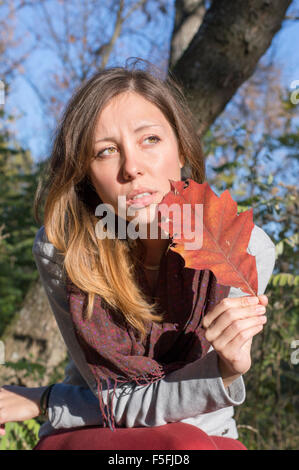 Sad girl holding red autumn leaf in her hand while sitting in the park Stock Photo