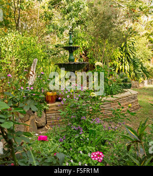 Decorative garden feature with low stone wall, ornate fountain, plants in containers, hemmed by lawn, flowers, shrubs, trees Stock Photo