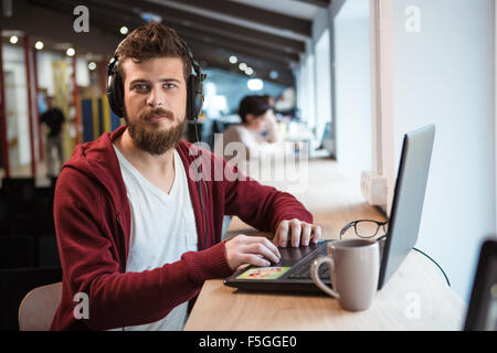 Young attractive hipster with beard sitting at the desk in earphones and working using laptop Stock Photo