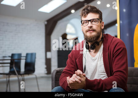 Hipster with beard in glasses and in brown hoodie sitting on sofa in the hall Stock Photo