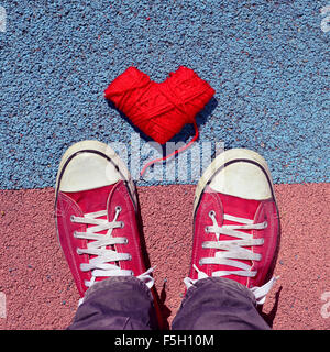 high-angle shot of a heart-shaped coil of red yarn and the feet of a man wearing red sneakers stepping on the asphalt Stock Photo