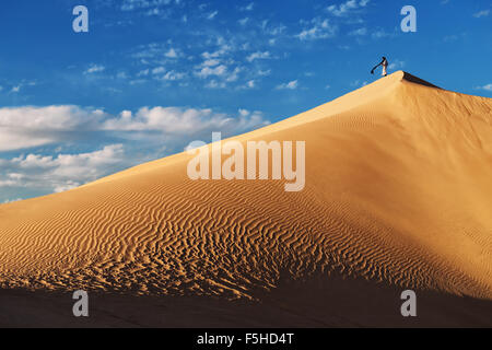 A traditional dressed Moroccan man stands on a sand dune against a cloudy blue sky. Stock Photo