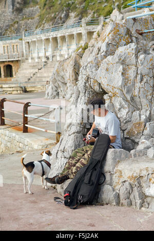 Man playing guitar with pet Jack Russell dog on rocks at sea-level by Plymouth Hoe, Devon, England Stock Photo