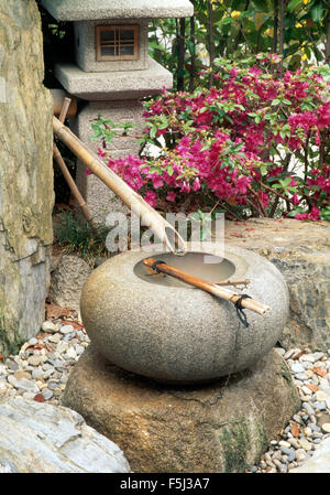 Close-up of a Japanese style bamboo pipe and circular stone water bowl beside a Japanese stone lantern Stock Photo