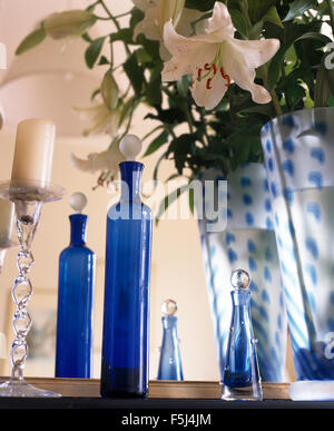 White lilies in blue glass vase on table with tall blue glass bottles and a clear glass candlestick Stock Photo