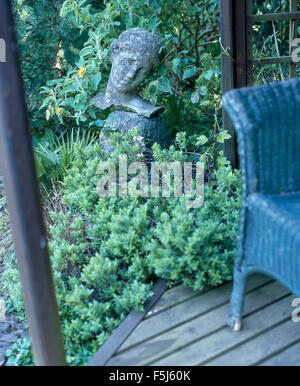 Blue painted wicker chair on decking beside a stone head under-planted with green plants in a summer garden Stock Photo