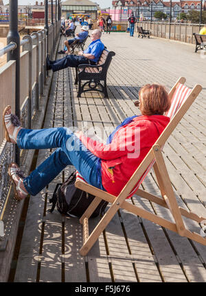 Person sitting on a deck chair and other people sat on benches at Skegness pier, Skegness, Lincolnshire, England, UK Stock Photo
