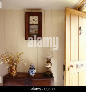 Antique wall clock on a painted tongue+groove paneled wall in a cottage hall with an Edwardian style lamp on a vintage chest Stock Photo