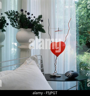 Red heart ornament on table in a nineties living room with a floral arrangement on a pedestal in front of white voile curtains Stock Photo