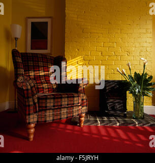 Red plaid upholstered armchair beside a small wood burning stove in bright yellow nineties living room with painted brick walls Stock Photo