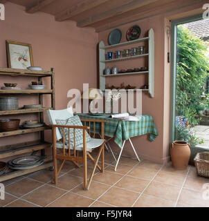 Canvas director's chair beside table with green checked cloth in a terracotta painted dining room with pottery on wooden shelves Stock Photo