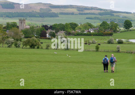 Couple of ramblers in beautiful countryside walking across a field towards rolling hills & scenic village of Gargrave (North Yorkshire, England, UK). Stock Photo