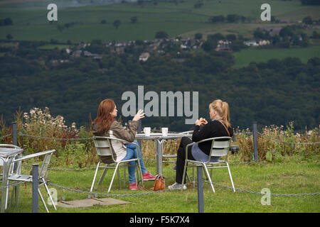 2 women friends sitting outside, eating & chatting at Cow & Calf Rock café, Ilkley, Yorkshire, UK - scenic spot with panoramic view over the valley. Stock Photo