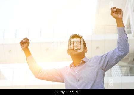 Successful Asian Indian businessman with arms up celebrating his victory, modern office building with morning sunlight as backgr Stock Photo