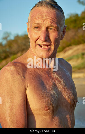 Mature aged man with blue eyes, walking on the beach just after a refreshing swim, water droplets all over him Stock Photo