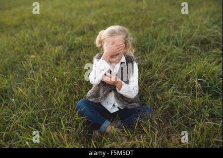 Young girl sitting on grass, covering eyes Stock Photo