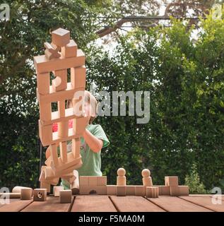 Young boy pushing over wooden structure made from building blocks, outdoors Stock Photo