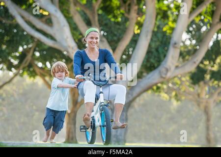 Mother sitting on son's bike, son pushing her along Stock Photo