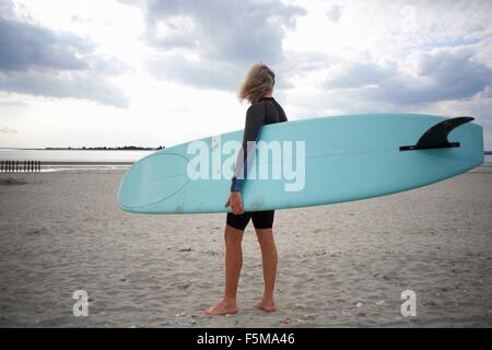 Senior woman standing on beach, holding surfboard, rear view Stock Photo