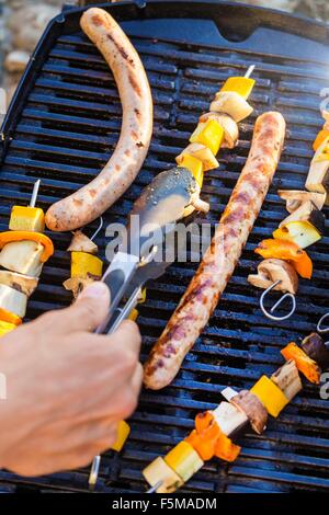 Person using tongs to turn sausages and kebabs on barbecue griddle Stock Photo