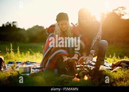 Young camping couple warming hands by campfire at dusk