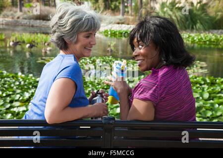 Two mature female friends sitting on park bench, laughing Stock Photo